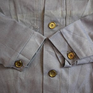 May include: Close-up of a light blue jacket with four front pockets and gold-colored buttons. The sleeves are cuffed, and the buttons are visible on the cuffs and front closure. The fabric has a subtle texture.