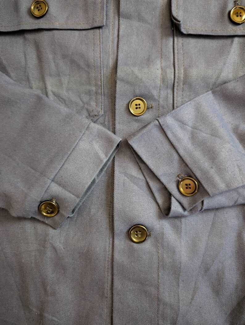 May include: Close-up of a light blue jacket with four front pockets and gold-colored buttons. The sleeves are cuffed, and the buttons are visible on the cuffs and front closure. The fabric has a subtle texture.