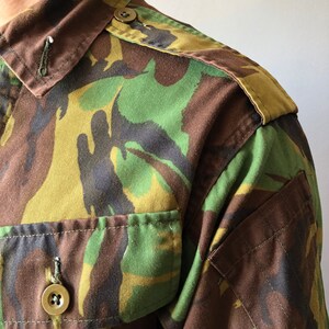 May include: A close-up of a green, brown, and black camouflage shirt with a buttoned collar and a patch on the shoulder.