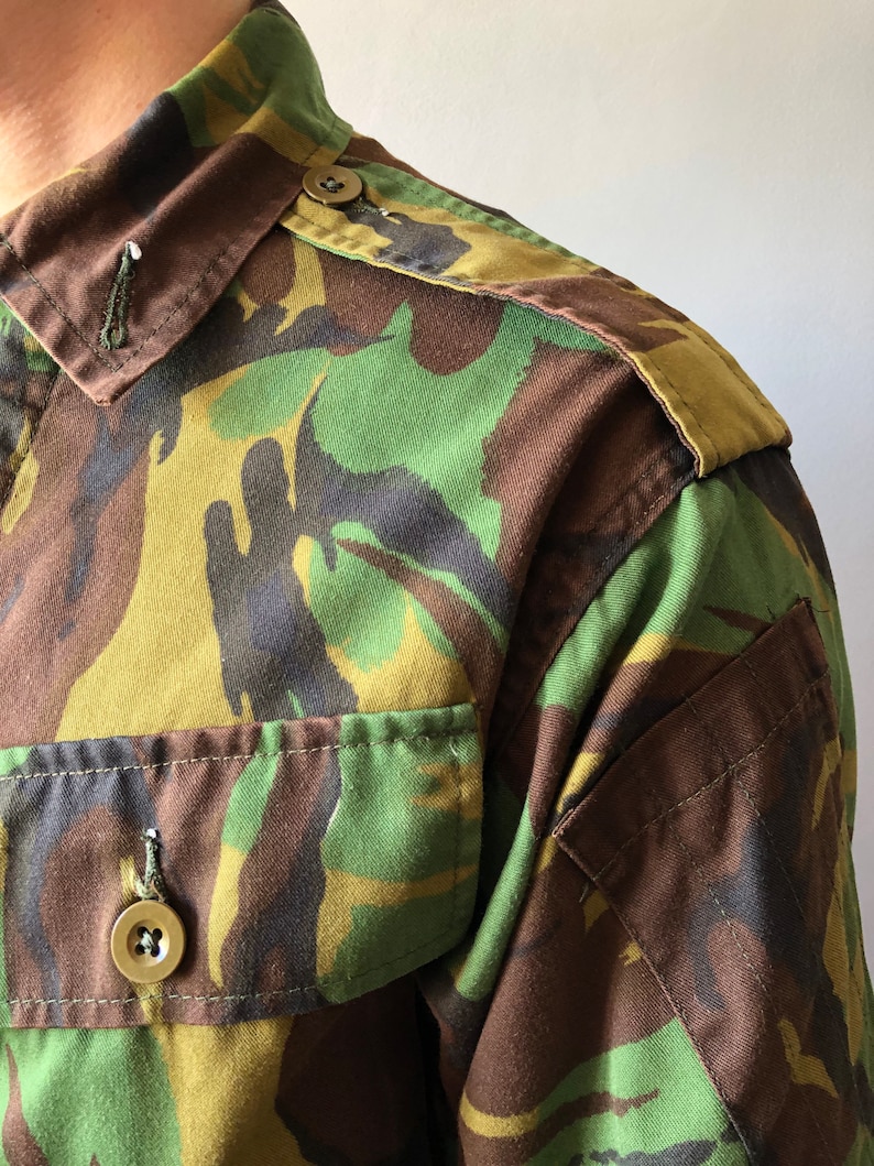 May include: A close-up of a green, brown, and black camouflage shirt with a buttoned collar and a patch on the shoulder.