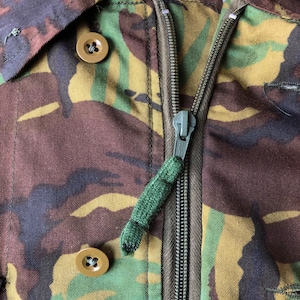 May include: Close-up of a camouflage jacket with a zipper and two buttons. The zipper has a green pull tab.