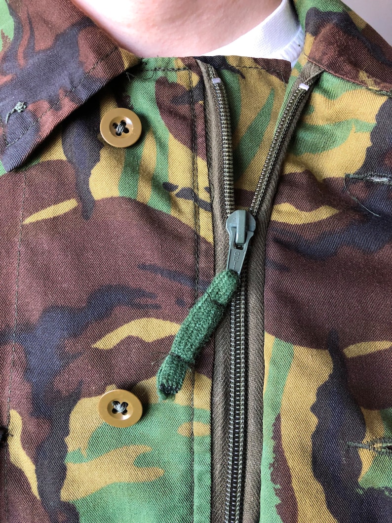 May include: Close-up of a camouflage jacket with a zipper and two buttons. The zipper has a green pull tab.
