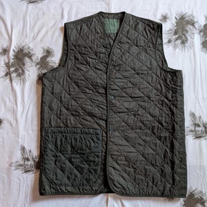 Könnte beinhalten: Eine dunkel olivgrüne, gesteppte Weste mit V-Ausschnitt und zwei Vordertaschen. Die Weste hat ein Rautenmuster und drei Knöpfe auf der Vorderseite. Die Weste liegt flach auf einer weißen, gemusterten Oberfläche.