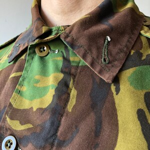 May include: Close-up of a camouflage fabric shirt with a buttoned collar. The fabric is a mix of green, brown, and black.