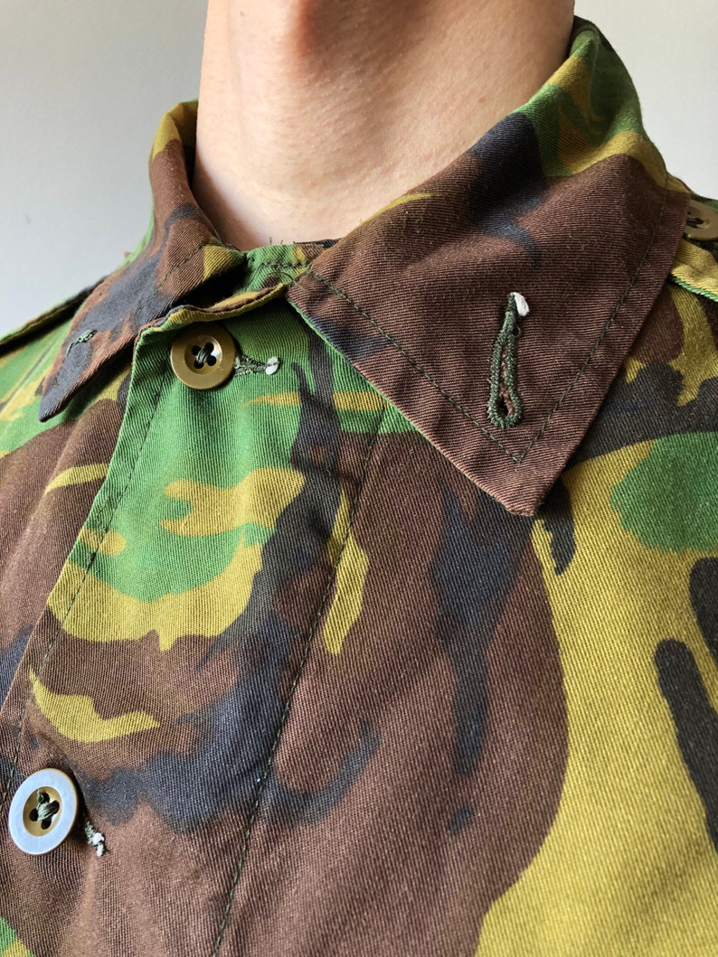 May include: Close-up of a camouflage fabric shirt with a buttoned collar. The fabric is a mix of green, brown, and black.