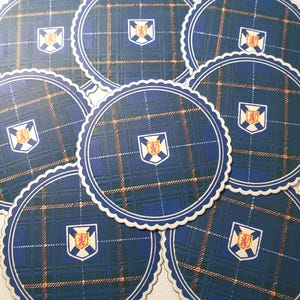 Peut inclure: Ensemble de dessous de verre ronds avec un motif écossais bleu et un bord festonné blanc. Chaque dessous de verre présente un blason au centre. Le motif écossais comprend des lignes bleu marine, orange et blanches.