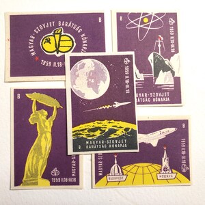 May include: Four purple matchboxes with yellow and white illustrations. The matchboxes feature the text "Magyar-Szovjet Baratsag Honapja" and the dates "1959 11.18-III.18". The illustrations include a rocket, a globe, a ship, and a building.