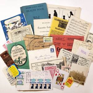 May include: A collection of vintage ephemera, including books, postcards, stamps, and documents. The items are arranged on a white surface, featuring a variety of colors and textures. Notable items include a blue health book and a playing card.