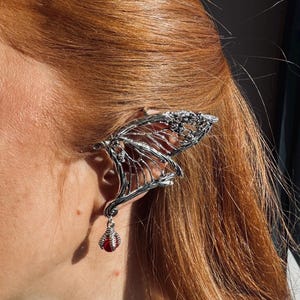 May include: A silver-toned ear cuff shaped like an intricate wing with a delicate, open-work structure. The piece features a dangling charm consisting of a small, dark red spherical bead held within a metallic claw-like setting at the base.