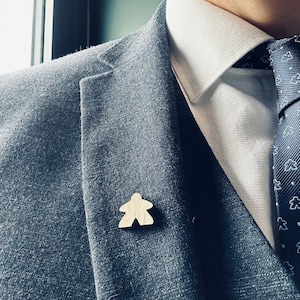 May include: A dark gray suit jacket with a white collared shirt and a dark blue patterned tie. A small wooden game piece is pinned to the lapel of the jacket.