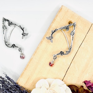 May include: Two silver ear cuffs with a floral and leaf design. Each cuff has a dangling pink crystal. The cuffs are displayed on a wooden surface with lavender and flowers.