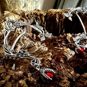 May include: A pair of silver ear cuffs with a floral design and red gemstone accents. The cuffs are shaped like branches with leaves and flowers, and the gemstones are set in claw settings.