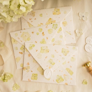 May include: Three handmade paper envelopes with a floral pattern in yellow and green, sealed with white wax seals. The envelopes are surrounded by white hydrangea flowers and a cream-colored ribbon.