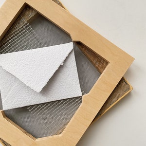 Könnte beinhalten: Eine Holzpapiermachform mit einem feinen Maschensieb, das einen weißen, strukturierten Papierumschlag hält. Die Form besteht aus hellfarbigem Holz und ist abgewinkelt, wodurch die geschichtete Konstruktion sichtbar wird. Die Szene spielt vor einem weißen Hintergrund.