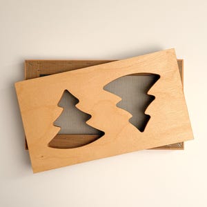 May include: A rectangular light-colored wooden board featuring two cutouts in the shape of evergreen trees. The board is positioned at an angle over a rectangular frame covered with a fine, dark-colored metal mesh screen.