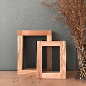 May include: Two rectangular wooden frames with a light brown natural wood grain finish, each featuring a fine dark mesh screen stretched across the center. The frames are positioned upright, with one slightly smaller frame placed in front of the larger one.
