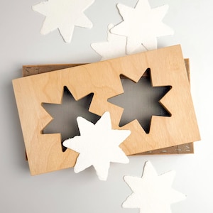 Pode incluir: Uma ferramenta de artesanato em madeira com dois recortes em forma de estrela, acompanhada por vários recortes de papel em forma de estrela brancos e texturizados. A ferramenta é castanho-claro, e as estrelas estão espalhadas numa superfície cinza-claro.