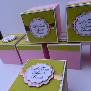 May include: A collection of pink and green gift boxes with gold detailing. Each box features a label with the text "A Toast Awaits Inside". The boxes are cube-shaped, with a textured green top and a pink body.