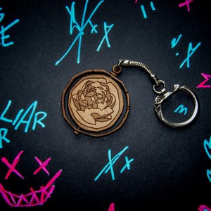 Op de afbeelding: Een houten sleutelhanger met een draaiend rond ontwerp met een bruine bloem. De sleutelhanger is bevestigd aan een zilveren metalen ring.