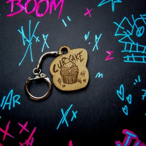 Op de afbeelding: Een houten cupcake-vormige sleutelhanger met een metalen ring en ketting. De sleutelhanger heeft het woord "CUPCAKE" en een cupcake-illustratie. De zwarte achtergrond is voorzien van graffiti-achtige tekeningen in roze en blauw.