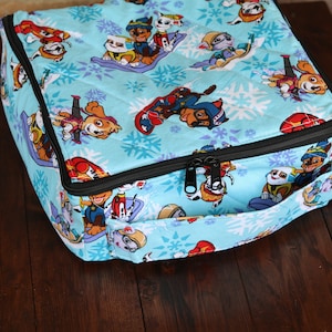 May include: A light blue, quilted fabric lunch bag featuring a repeating pattern of cartoon dogs on snowboards and snowflakes. The bag has a black zipper closure and a small handle on one side.