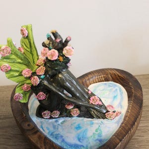 May include: A dark-toned fairy sculpture with pink roses and green leaves, seated in a heart-shaped wooden bowl. The fairy has a floral crown and wings. The bowl's interior features a blue and white marbled design.