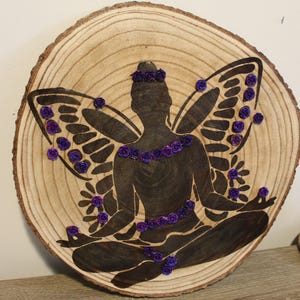 May include: A wooden slice with a dark silhouette of a fairy with butterfly wings. The fairy is adorned with purple rose accents on the wings, crown, and necklace. The wood has a natural grain pattern.