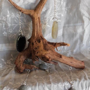 May include: A rustic wooden jewelry display stand with a natural, textured appearance. The stand features multiple silver chains with various pendant-style earrings in black, gray, and yellow-green. The stand is set against a soft, patterned fabric.