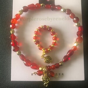 May include: A set of two red and gold beaded bracelets. The larger bracelet has a charm with a gold rose. The smaller bracelet is a ring with a gold rose charm.