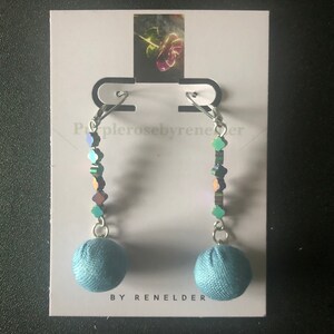 May include: A pair of blue fabric ball earrings with iridescent green and purple beads hanging from silver hooks. The earrings are on a white card with the text "Purple rose by renelder" and "By ReneLder".