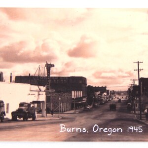 May include: A black and white vintage photograph of a street scene in Burns, Oregon in 1945. The photo shows a street lined with buildings, including a hotel, and cars parked along the side of the road.