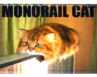 May include: A humorous image of a cat with orange fur, looking intently at the camera, with its mouth open as if it is about to bite a black metal bar. The text "MONORAIL CAT" is printed in black letters at the top of the image.