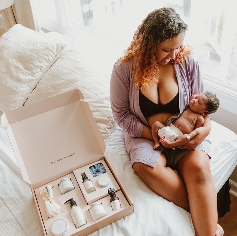 May include: A pink and white gift box with the text "Find beauty in the moment."  The box is open and contains a variety of bath and body products, including a bottle of lotion, a bottle of body wash, a bar of soap, and a jar of bath salts. A woman is sitting on a bed holding a baby.