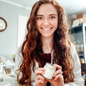 May include: A woman with long, curly red hair smiles while holding a small white jar with a silver lid. The jar is filled with a white cream.