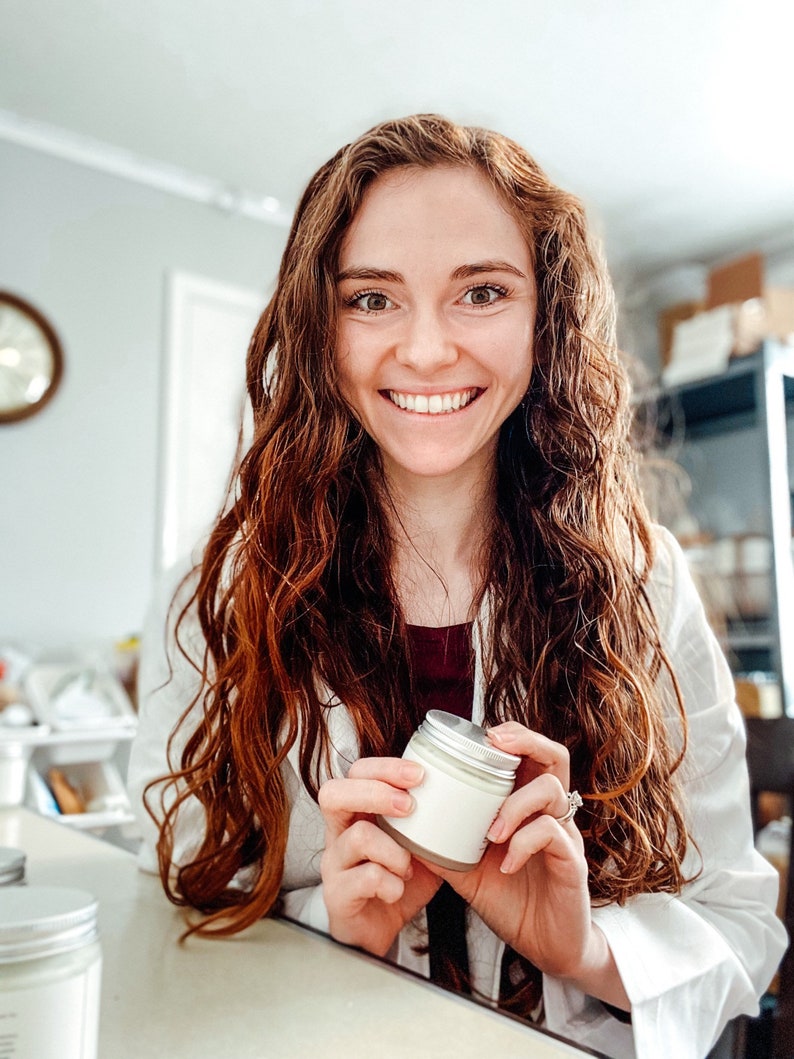 May include: A woman with long, curly red hair smiles while holding a small white jar with a silver lid. The jar is filled with a white cream.