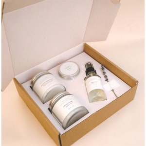 May include: A gift box set containing three candles and a bottle of body spray. The candles are in glass jars with silver lids and labels that say "Lavender & Honey" and "Calming Lavender". The body spray bottle is clear glass with a silver lid and a label that says "Calming Lavender Body Mist".