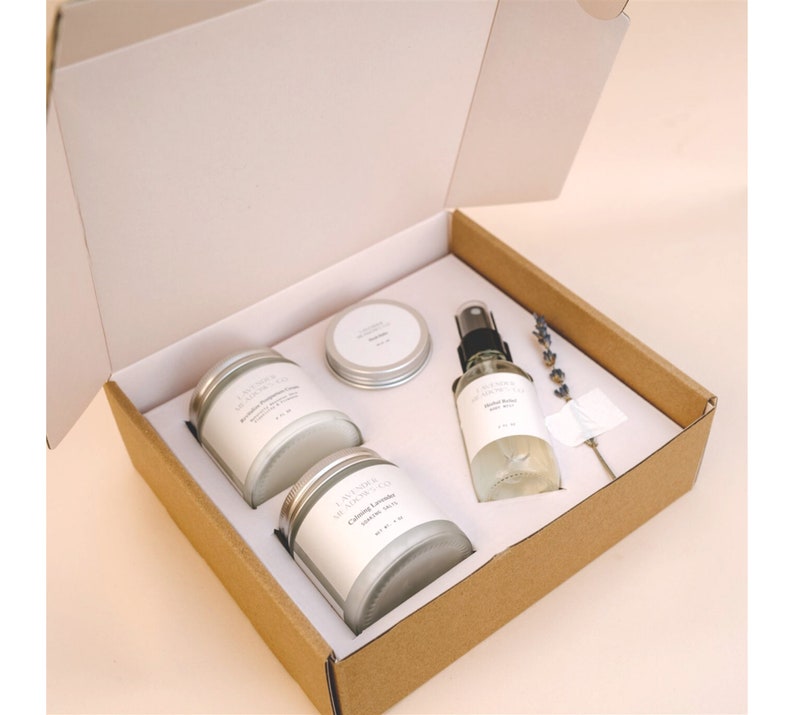 May include: A gift box set containing three candles and a bottle of body spray. The candles are in glass jars with silver lids and labels that say "Lavender & Honey" and "Calming Lavender". The body spray bottle is clear glass with a silver lid and a label that says "Calming Lavender Body Mist".