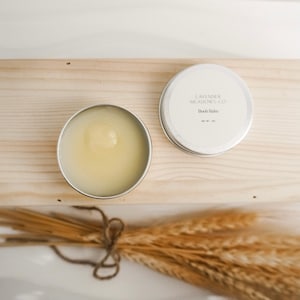 May include: A silver tin with a white label that reads "Lavender Meadows Co.  Body Balm" and a net weight of 1 oz. The tin is open and the balm is a light yellow color. The tin is sitting on a wooden surface with a bundle of wheat stalks in the foreground.