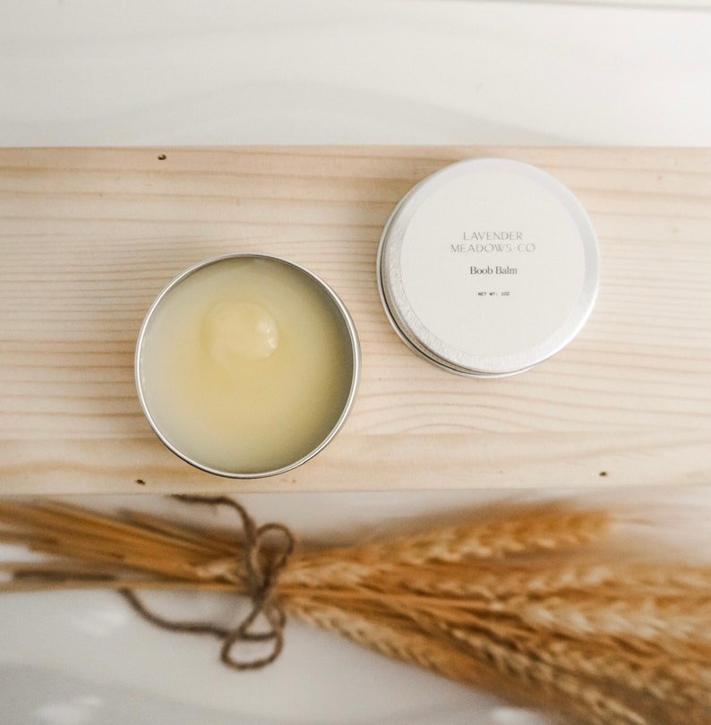 May include: A silver tin with a white label that reads "Lavender Meadows Co.  Body Balm" and a net weight of 1 oz. The tin is open and the balm is a light yellow color. The tin is sitting on a wooden surface with a bundle of wheat stalks in the foreground.