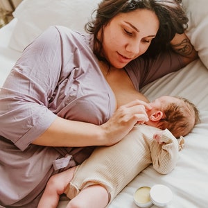 May include: A mother in a purple robe breastfeeds a baby in a cream onesie on a white bed. Two small round containers of balm are on the bed.