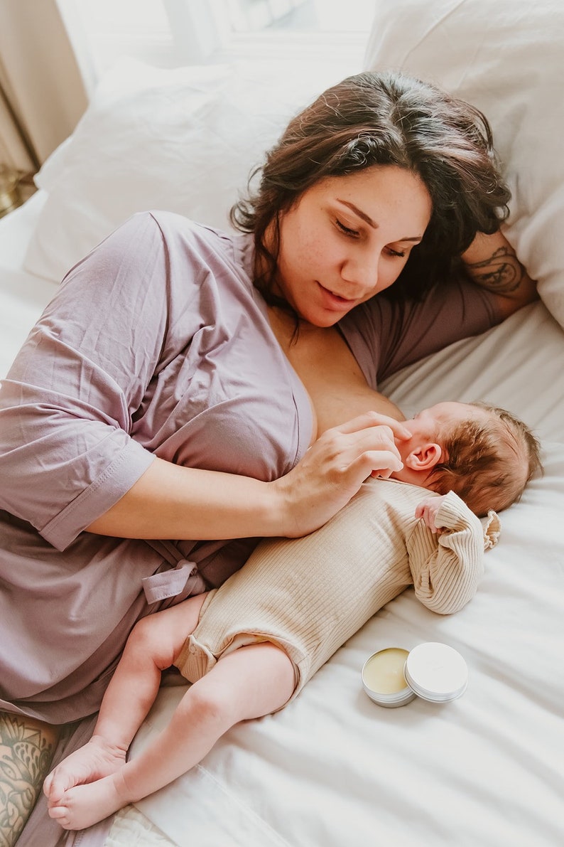 May include: A mother in a purple robe breastfeeds a baby in a cream onesie on a white bed. Two small round containers of balm are on the bed.