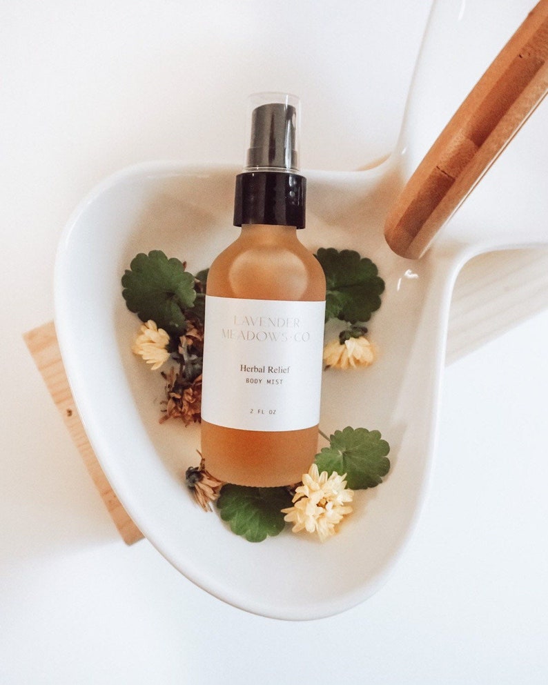 May include: A clear glass bottle with a black spray top filled with a light amber liquid. The bottle is labeled "Lavender Meadow Co. Herbal Relief Body Mist 2 fl. oz.". The bottle is surrounded by small white flowers and green leaves.
