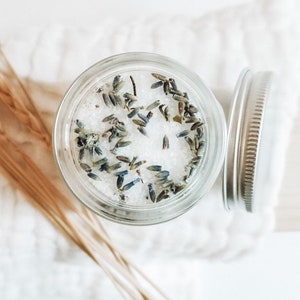 May include: A glass jar filled with white bath salts and dried lavender flowers. The jar has a silver lid.