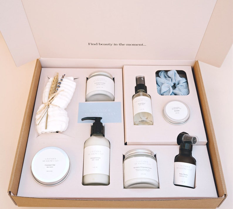 May include: A gift box filled with lavender scented bath and body products. The box includes a white washcloth, a blue hair scrunchie, a bottle of lavender body spray, a jar of coconut oil, a bottle of lavender body lotion, a jar of lavender body cream, and a bottle of baby oil.