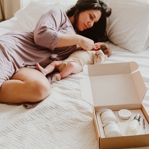 May include: A mother and baby lying on a white bed, with an open box containing a set of bath products. The box contains three white jars and a bottle with a spray nozzle. The products are arranged in a neat row.