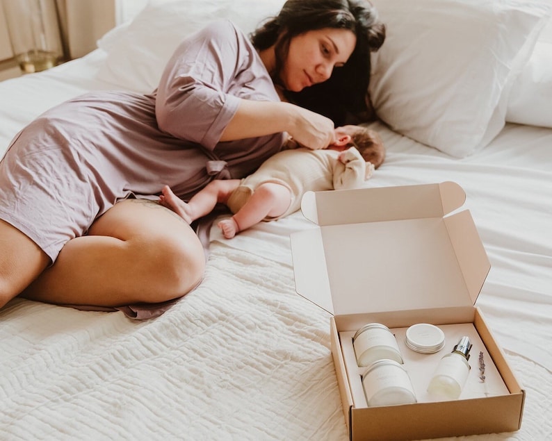 May include: A mother and baby lying on a white bed, with an open box containing a set of bath products. The box contains three white jars and a bottle with a spray nozzle. The products are arranged in a neat row.