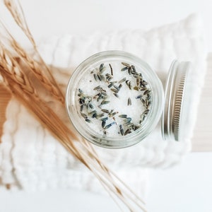 May include: A glass jar filled with white bath salts and dried lavender flowers. The jar has a silver lid and is sitting on a white surface.