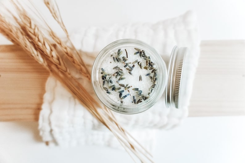 May include: A glass jar filled with white bath salts and dried lavender flowers. The jar has a silver lid and is sitting on a white surface.