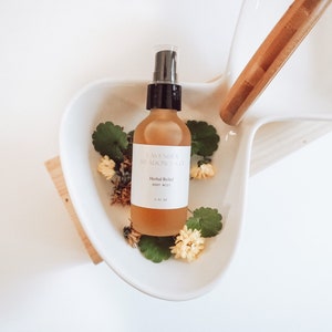 May include: A clear glass bottle with a black spray top filled with a light brown liquid. The bottle is labeled "Lavender Meadow Co. Herbal Relief Body Mist 118 ml". The bottle is sitting in a white ceramic bowl with green leaves and yellow flowers.