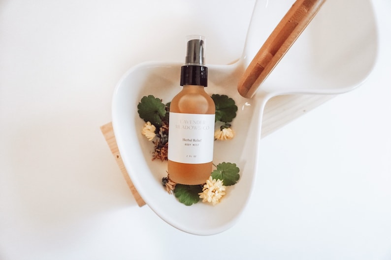 May include: A clear glass bottle with a black spray top filled with a light brown liquid. The bottle is labeled "Lavender Meadow Co. Herbal Relief Body Mist 118 ml". The bottle is sitting in a white ceramic bowl with green leaves and yellow flowers.